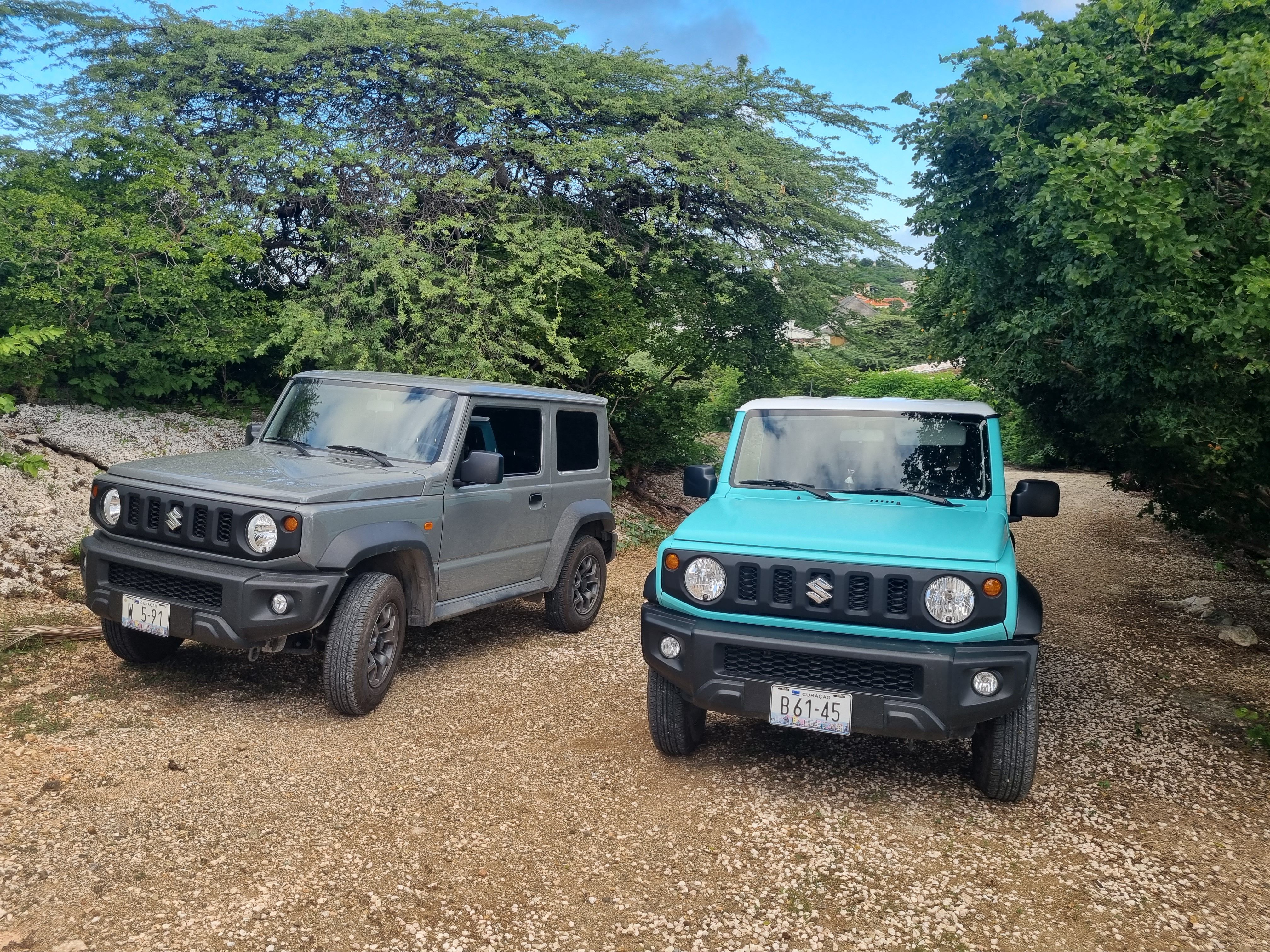 A grey and a turquoise Suzuki Jimny SUV parked side-by-side on a dirt path.