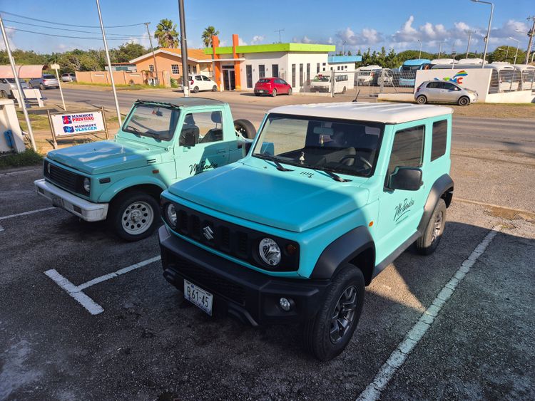 Two turquoise Suzuki Jimny SUVs, an old and new model, parked side-by-side outdoors.