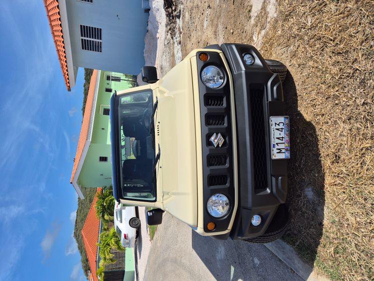 Front view of a tan Suzuki Jimny with a black grille parked outside under a clear sky.