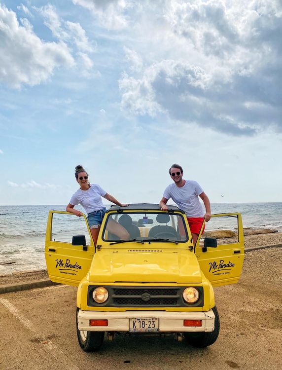 A smiling couple stands beside a yellow jeep on a beach.