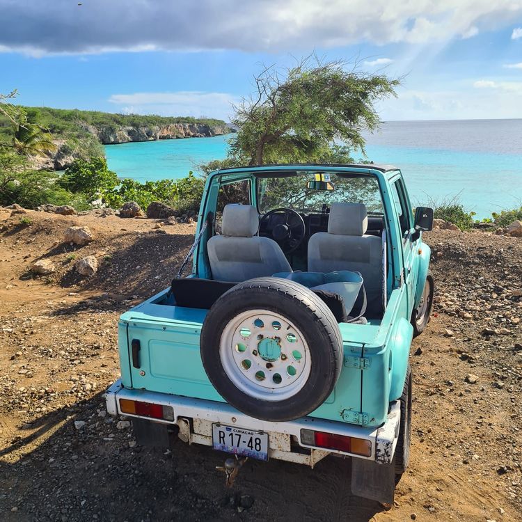 Teal open-top SUV parked on a dirt road overlooking a turquoise ocean and cliffs.