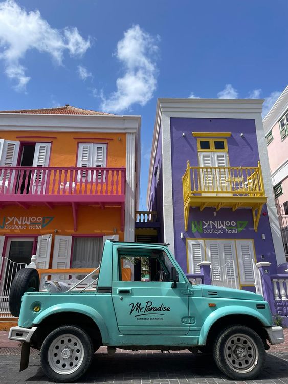 Turquoise vintage jeep parked in front of vibrant orange and purple buildings.