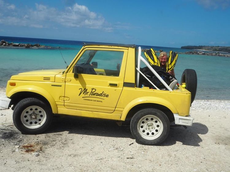 Yellow jeep parked on a sandy beach with diving gear near turquoise water.