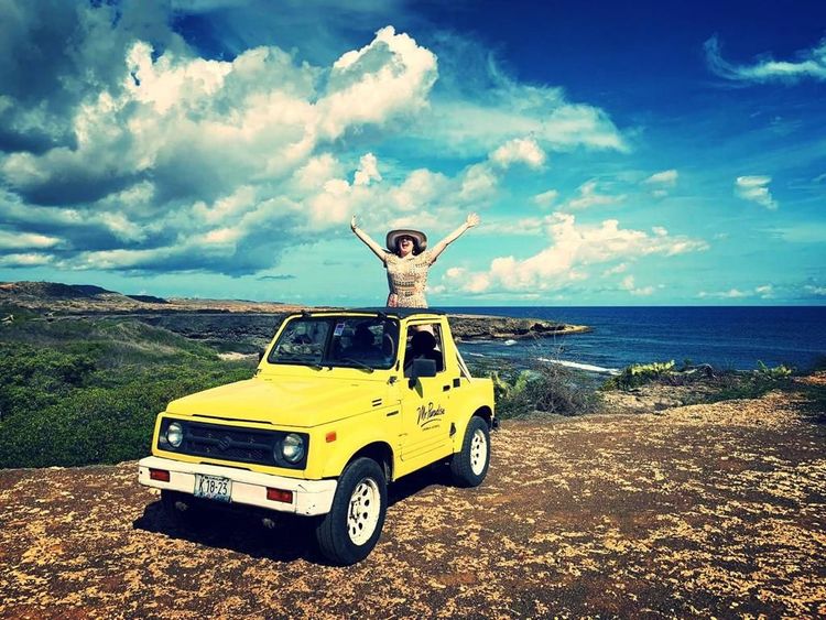Woman joyfully stands in a yellow jeep overlooking a beautiful ocean landscape.