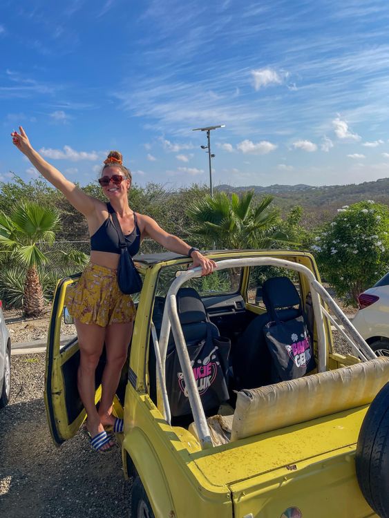 Joyful woman in sunglasses stands in a yellow open-top jeep on a sunny day.