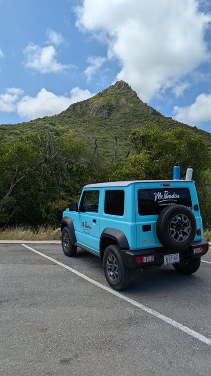 Light blue Suzuki Jimny SUV parked on asphalt before a large, green, conical mountain.