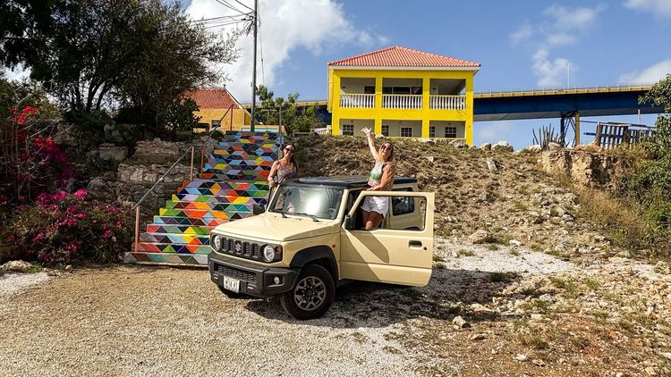 Two women wave from a beige Suzuki Jimny beside colorful mosaic stairs and a yellow house.