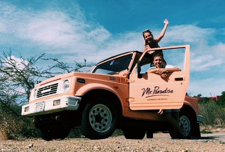 Two happy people in a peach open-top off-road vehicle marked 'Mr. Paradise' in arid landscape.