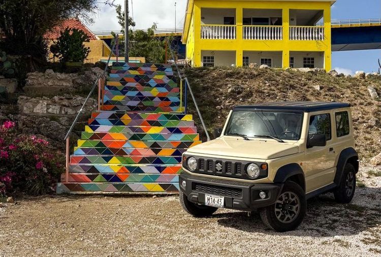 Beige Suzuki Jimny by a vibrant geometric staircase leading to a yellow building.